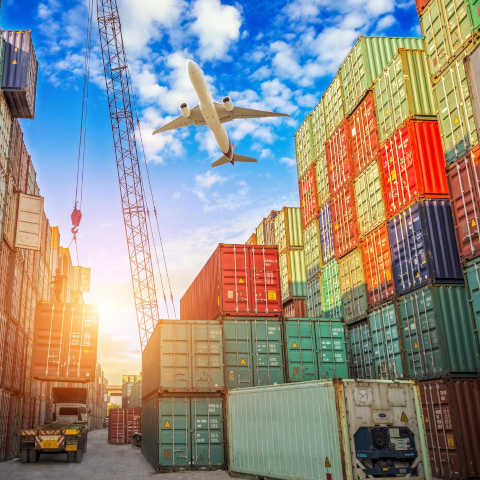 busy shipping area containing shipping containers, a forklift and an airplane flying overhead