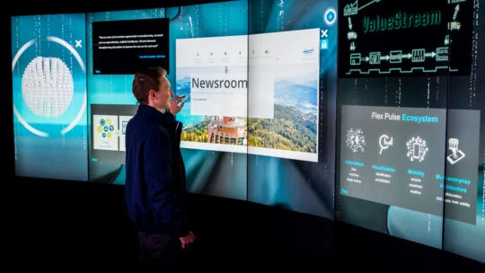 Person interacting with a large curved wall of illuminated digital displays showing dashboards, diagrams, and a newsroom webpage in a high‑tech environment.