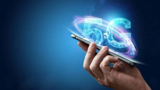 Hand holding a smartphone with a glowing blue “5G” graphic swirling above it against a dark blue futuristic background.