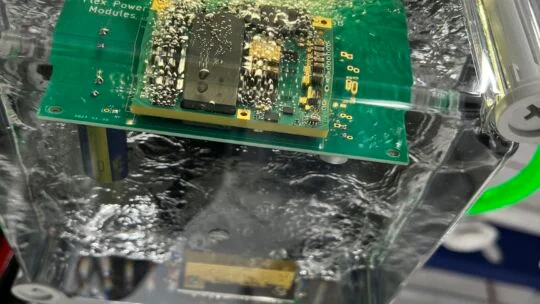 A close‑up view of an electronics cooling test setup showing a Flex Power Modules circuit board submerged in a liquid cooling chamber, with bubbles forming around the components during thermal evaluation.