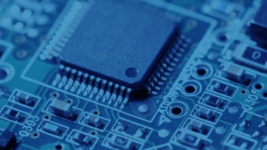 Close‑up of a blue‑tinted circuit board featuring a central integrated chip surrounded by fine soldered pins, traces, and various electronic components.