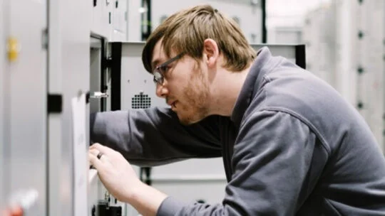 Técnico que trabaja dentro de un centro de datos, inspeccionando y ajustando componentes dentro de un gabinete eléctrico o de servidores de gran tamaño.
