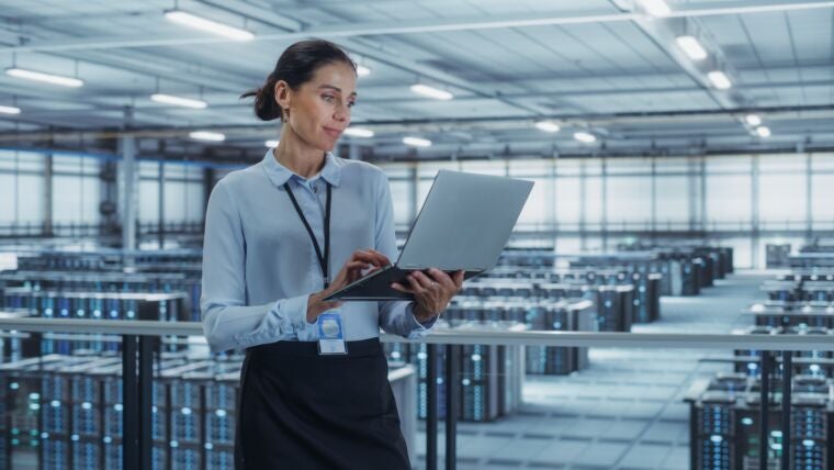 Person standing in a large data center holding an open laptop and working while surrounded by rows of server racks optimized for networking for AI data centers